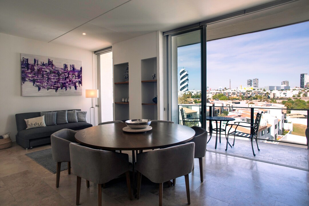 A spacious living and dining area is enhanced by large glass doors that provide access to a private balcony. The round dining table is surrounded by gray upholstered chairs. A minimalist artwork adorns the wall, and a comfortable sofa is positioned nearby.