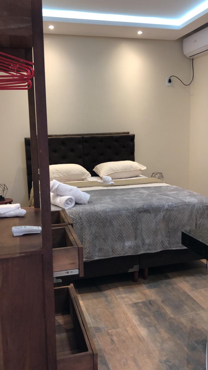 The room features a dark upholstered headboard positioned above a neatly arranged bed with white pillows. Soft gray bedding complements the decor. A wooden drawer unit and towels are neatly organized nearby. Warm light fixtures illuminate the space, enhancing its inviting ambiance.