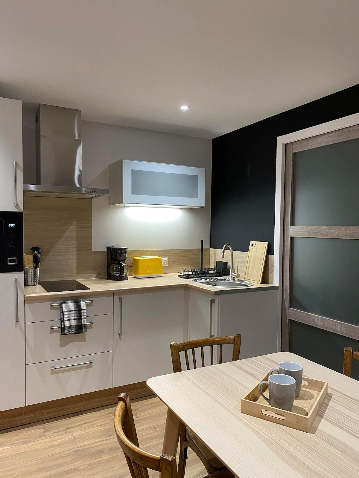 A modern kitchen area is equipped with sleek cabinetry and stainless-steel appliances. A small dining table with two wooden chairs is set, featuring a tray with cups. A coffee maker and yellow toaster are visible, providing essential kitchen amenities.