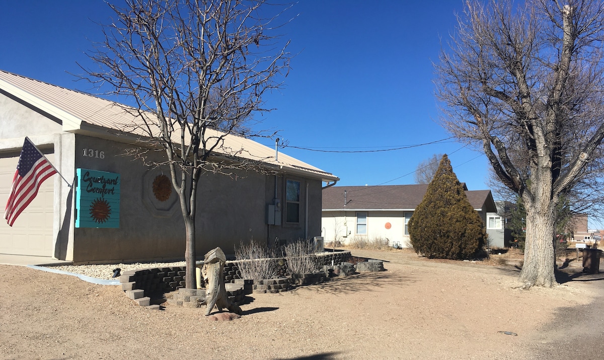 The image captures a quiet street scene featuring a single-story building adorned with colorful sun motifs. A large tree and smaller shrub provide natural elements to the landscape, with a flag gently waving. The unpaved ground leads to several nearby structures.