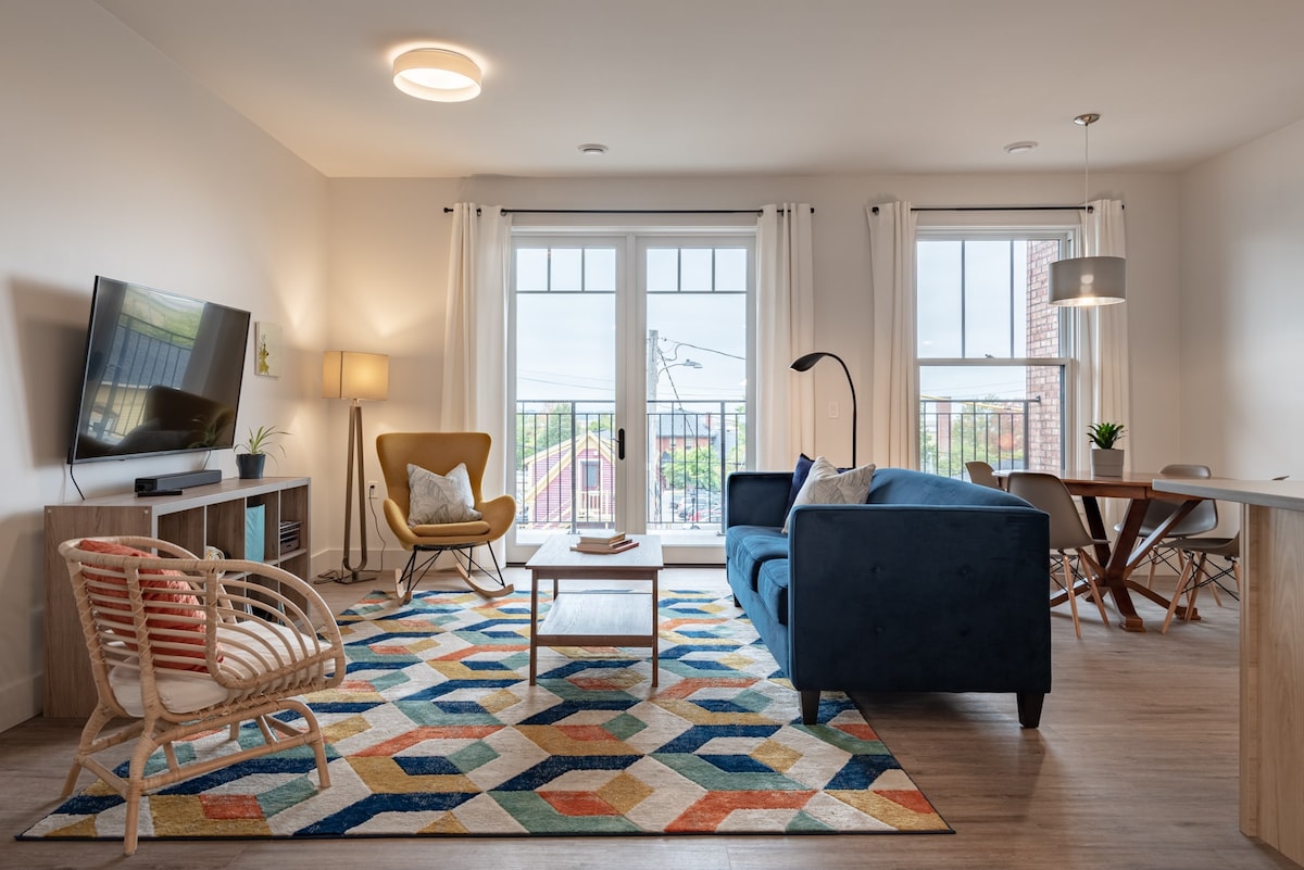 The living area features a blue sofa, a yellow accent chair, and a stylish table with four seats. A large TV is mounted on the wall, with a soft rug below that complements the decor. Large windows allow natural light, offering views of the surroundings.