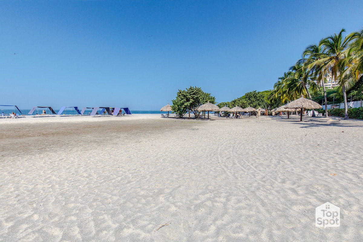 A wide sandy beach is depicted with a clear blue sky overhead. Umbrellas and shaded seating areas are scattered along the shore, providing spaces for relaxation. Lush palm trees are visible on the right, contributing to the inviting coastal atmosphere.