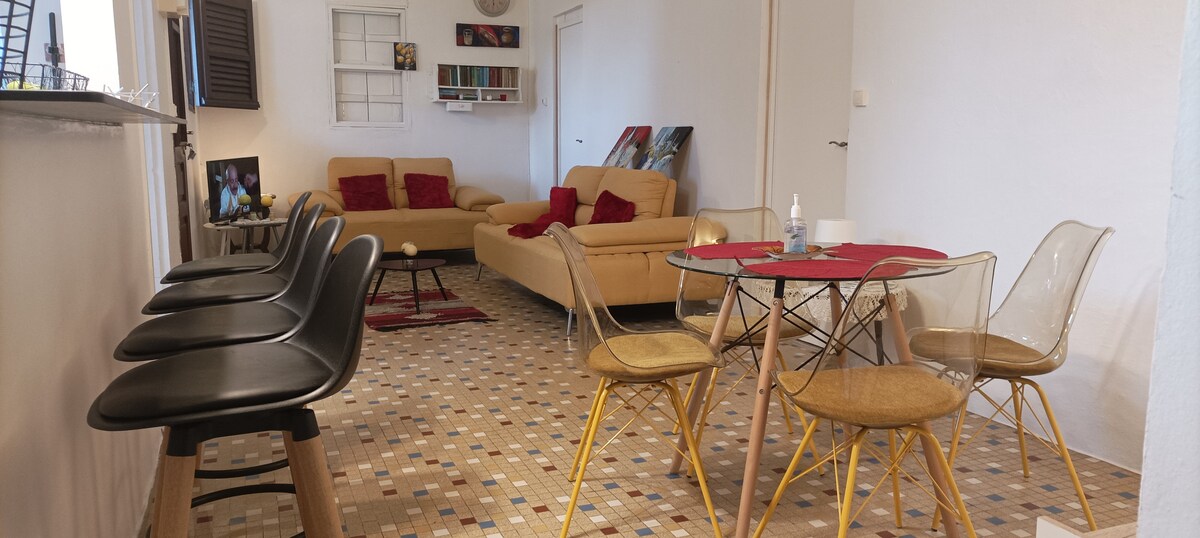 An open living space features a light-colored sectional sofa, complemented by two red cushions. A round dining table surrounded by transparent chairs occupies one side, while a mosaic floor adds visual interest. Decorative items and artworks are visible on the walls.