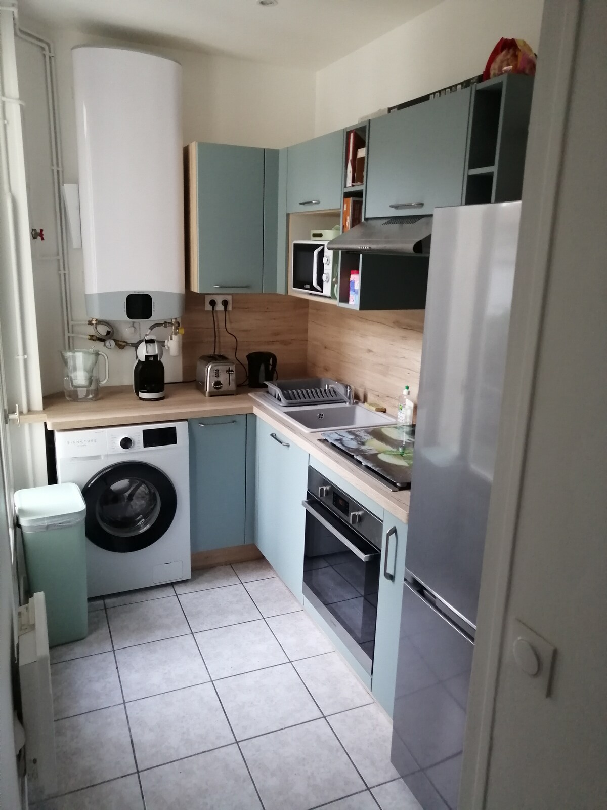 A compact kitchen features modern cabinetry in light blue and natural wood tones. Appliances include a washing machine, oven, and refrigerator, with a microwave positioned above the counter. A sink and stove are integrated into the workspace, and tiles cover the floor for easy maintenance.