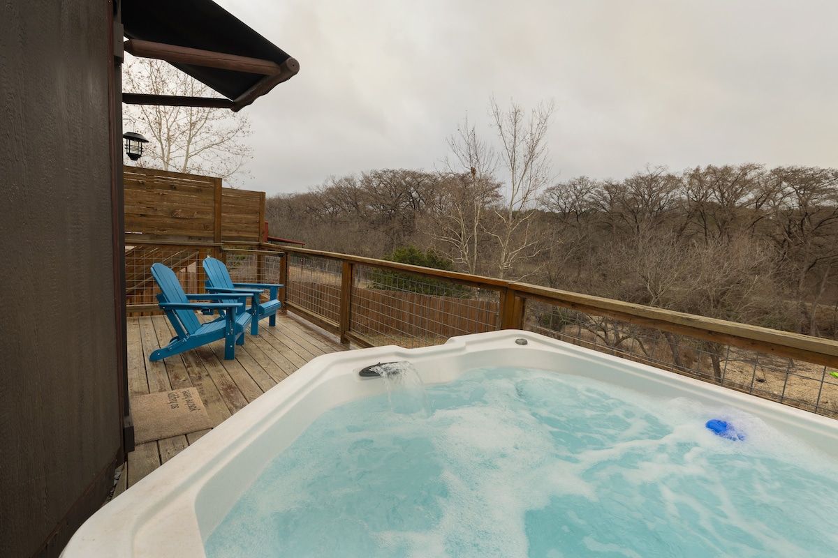 A private hot tub is positioned on the deck, offering serene views of the surrounding landscape. Two blue Adirondack chairs are placed nearby, complemented by a wooden railing and decking that enhance the outdoor relaxation space.
