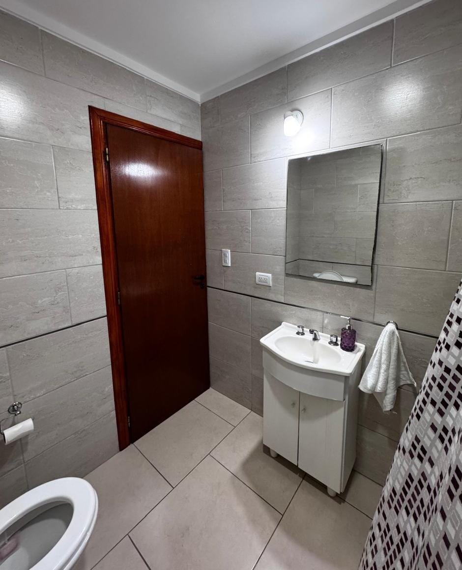 The bathroom features neutral-colored tiles covering the walls and floor. A compact sink is situated adjacent to a tall mirror, with a towel hanging nearby. A wooden door provides access, while a shower with a patterned curtain is visible, creating a clean and functional space.