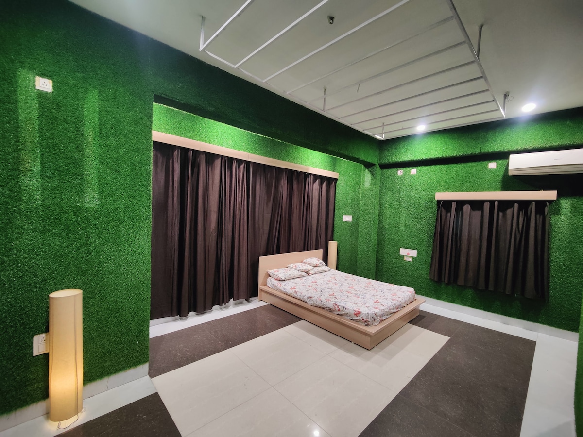 A spacious sleeping area features a bed with a floral-patterned blanket against a backdrop of vibrant green grass-like walls. Dark curtains frame two windows, and modern lamps illuminate the corners of the room, complementing the sleek tile flooring.