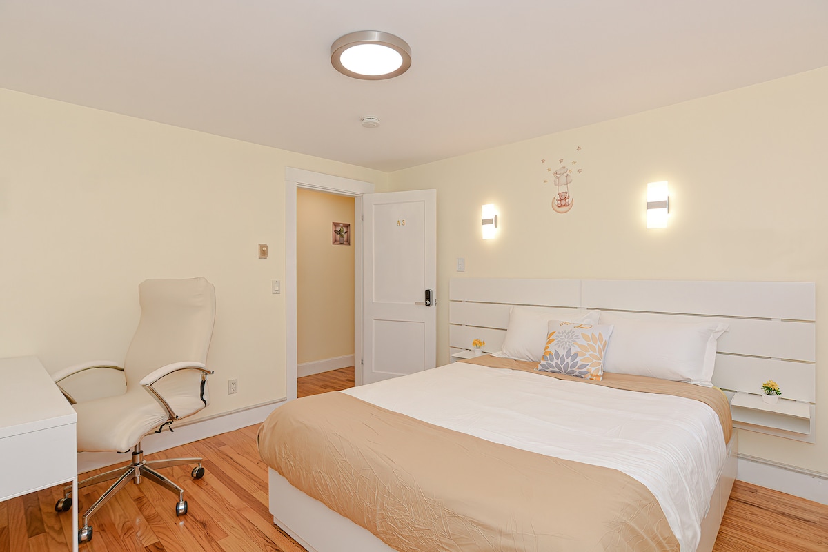 A modern bedroom features a spacious bed with neutral-toned bedding. A dedicated workspace is visible, equipped with a rolling chair and desk. Soft lighting is provided by wall-mounted fixtures, and a door leads to the hallway, indicating easy access.