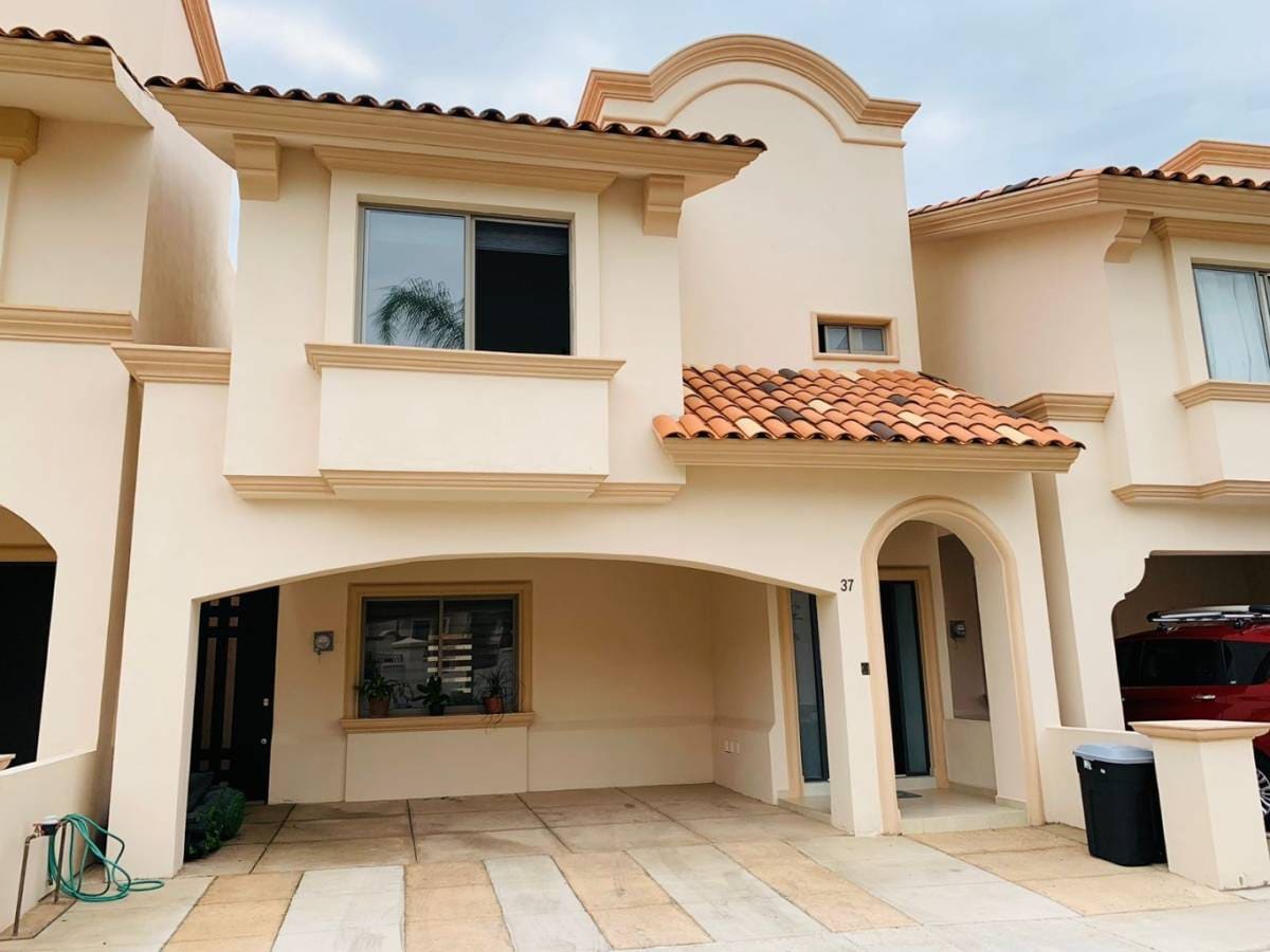 A two-story building features a terracotta-tiled roof and soft beige façade. The entrance is highlighted by an archway and large windows, bringing in natural light. An outdoor area with paved stone is visible, providing space for relaxation or gatherings.
