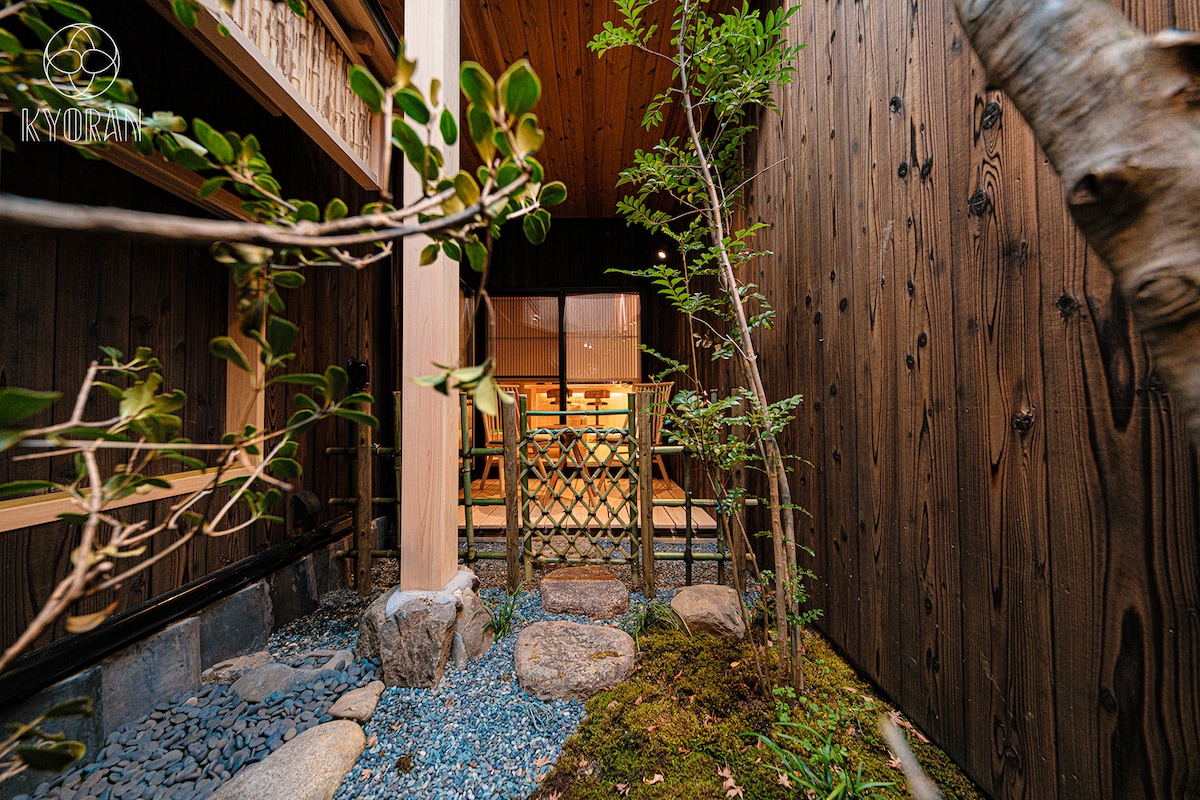 The image captures a serene courtyard featuring natural stone pathways and green foliage. A wooden fence offers gentle separation from the inviting interior visible through large windows. Subtle landscaping elements, including rocks and small plants, enhance the tranquil atmosphere of the space.