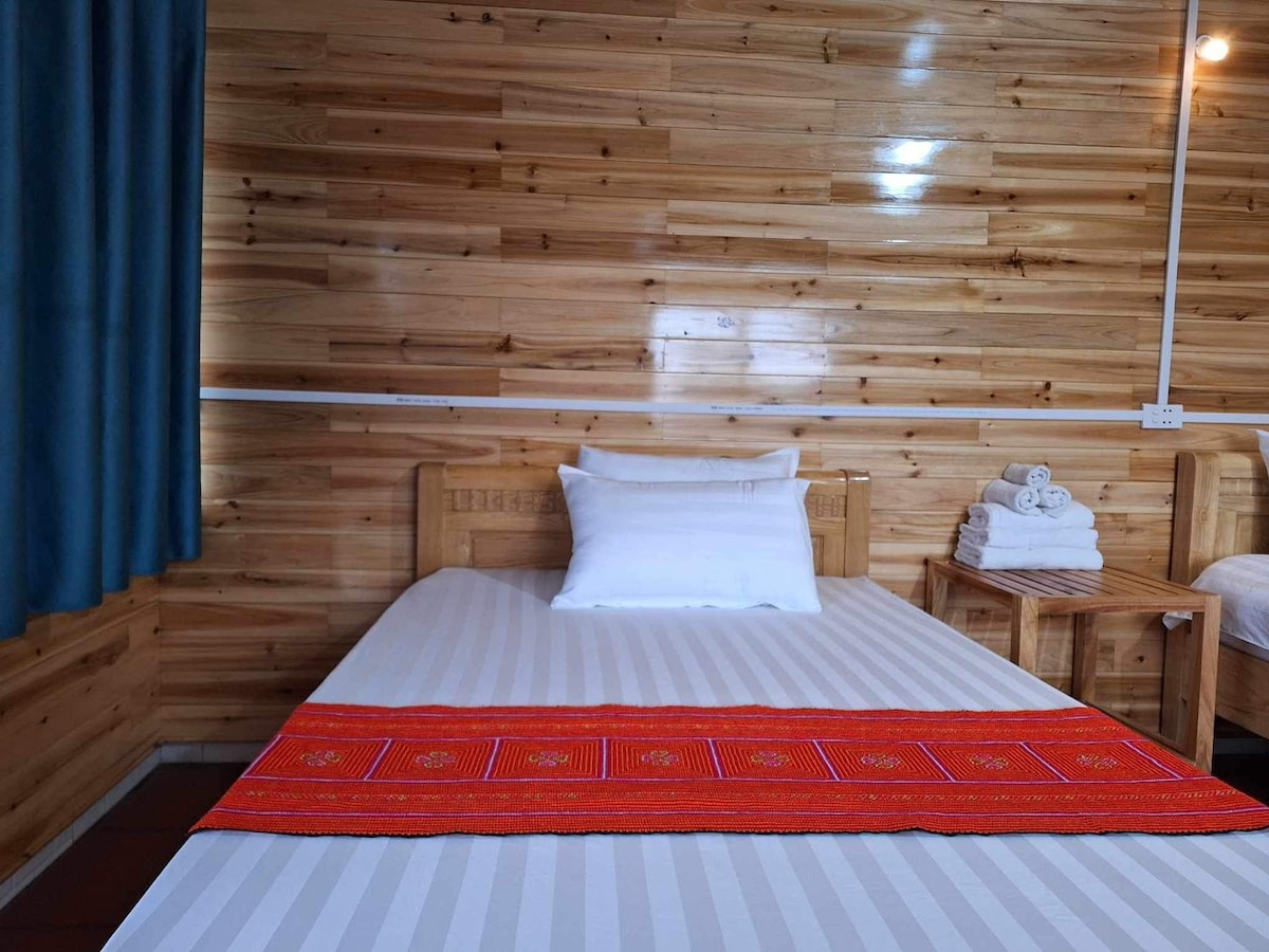 A spacious bed is adorned with a vibrant red runner, and a set of clean towels is neatly arranged on a side table. Light filters through a window dressed with blue curtains, illuminating the wooden walls of the room.