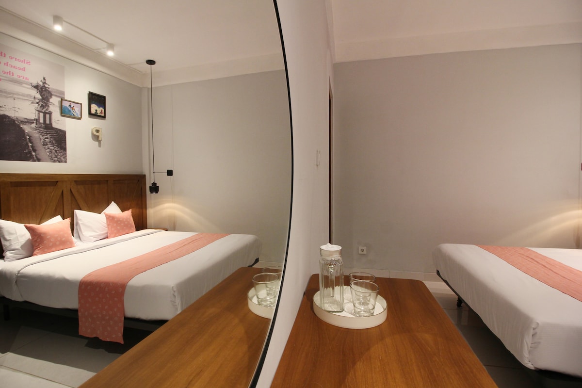 A bright and modern room features soft grey walls and a spacious bed adorned with a pink runner. A wooden bedside table holds a glass jug and two glasses. A large mirror reflects the bed and adds depth to the space, enhancing the overall ambiance.