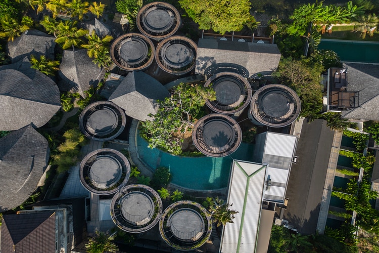 An aerial view captures the unique circular structures of the property, surrounded by lush greenery. A central swimming pool reflects the natural beauty, while various rooftops and pathways are visible, illustrating the thoughtful layout and design of the accommodation.