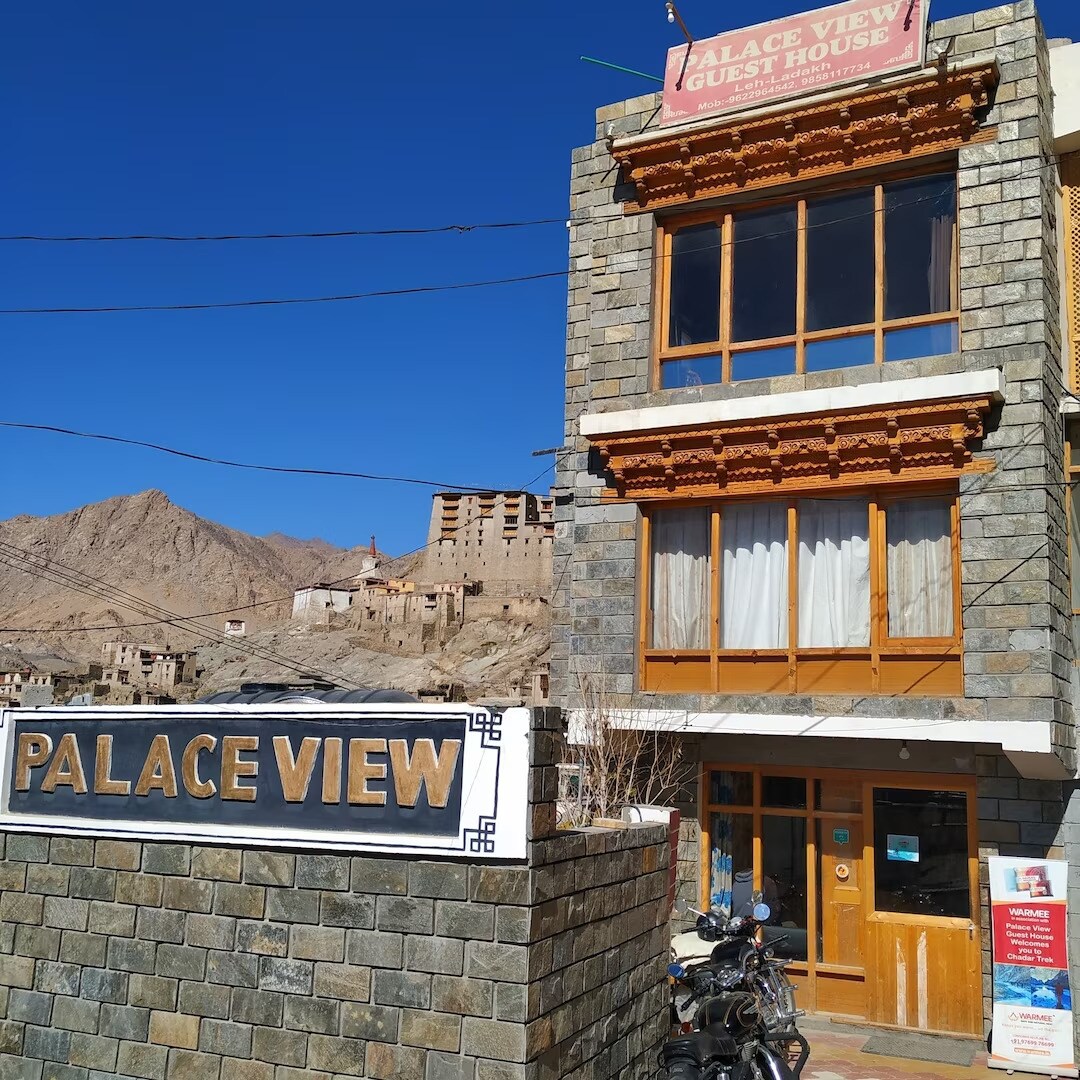 The exterior of the guest house features a stone facade with wooden accents and multiple large windows. A sign reading 'Palace View Guest House' is prominently displayed above the entrance. The surrounding mountainous landscape is visible against a clear blue sky.