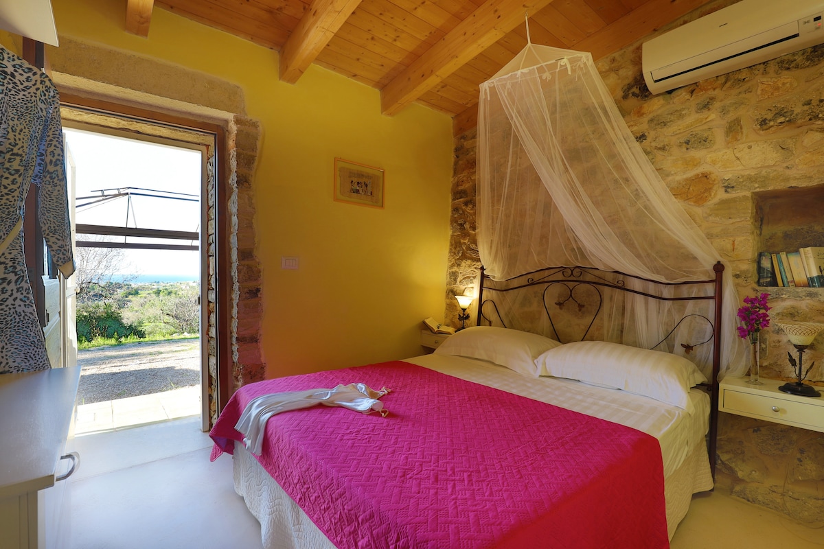 A cozy bedroom features a double bed draped with a netting canopy. The walls are adorned with warm yellow tones, and stone elements add character. Natural light enters from a door leading to a terrace, offering scenic views of the surrounding landscape.