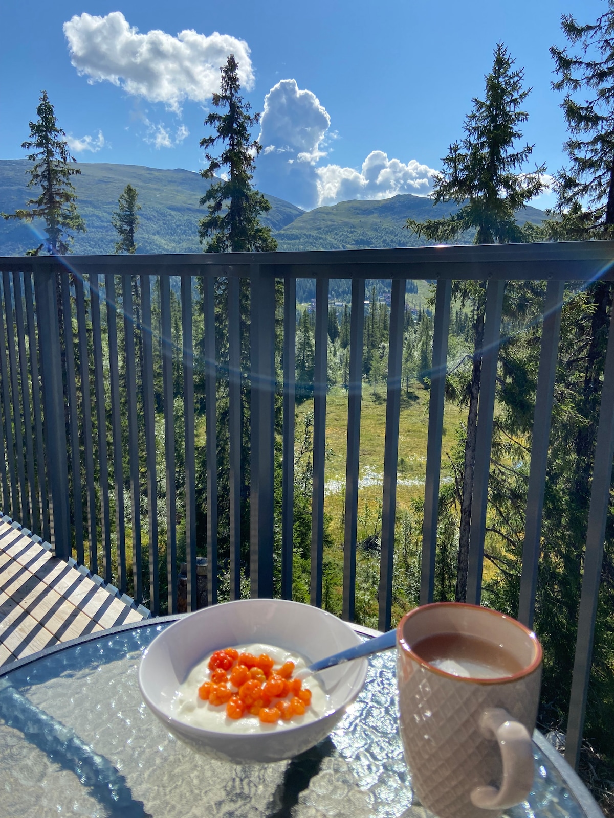 A dining table on a balcony overlooks a scenic view of rolling hills and trees, under a bright blue sky. A bowl of yogurt topped with orange berries and a cup of coffee are positioned on the glass surface, inviting a peaceful breakfast experience.