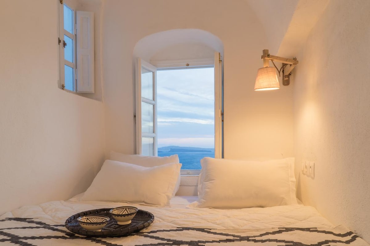 A serene sleeping area features a spacious bed adorned with white linens. Natural light enters through an arched window, which frames a view of the sea. A wall-mounted lamp offers soft illumination, and decorative plates are placed on a woven tray nearby.