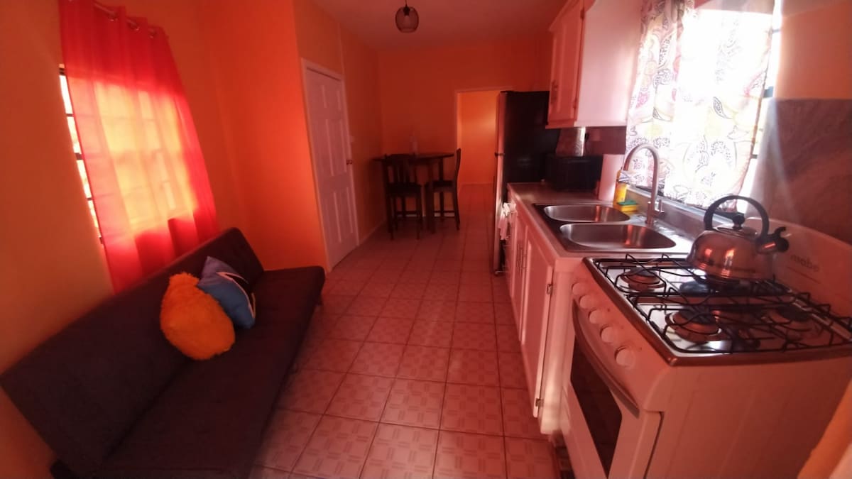 A well-lit kitchen area features a gas stove, sink, and refrigerator set against bright orange walls. A cozy seating area with a futon and colorful cushions is visible. Natural light streams through the window, adorned with bright curtains, while a small dining space is positioned in the background.