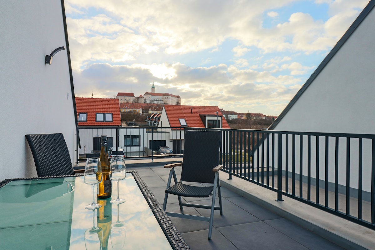 A rooftop terrace features a glass table set with three wine glasses and a bottle. Lounge chairs are positioned nearby, offering a view over a landscape of rooftops against a cloudy sky, with a castle visible in the distance.