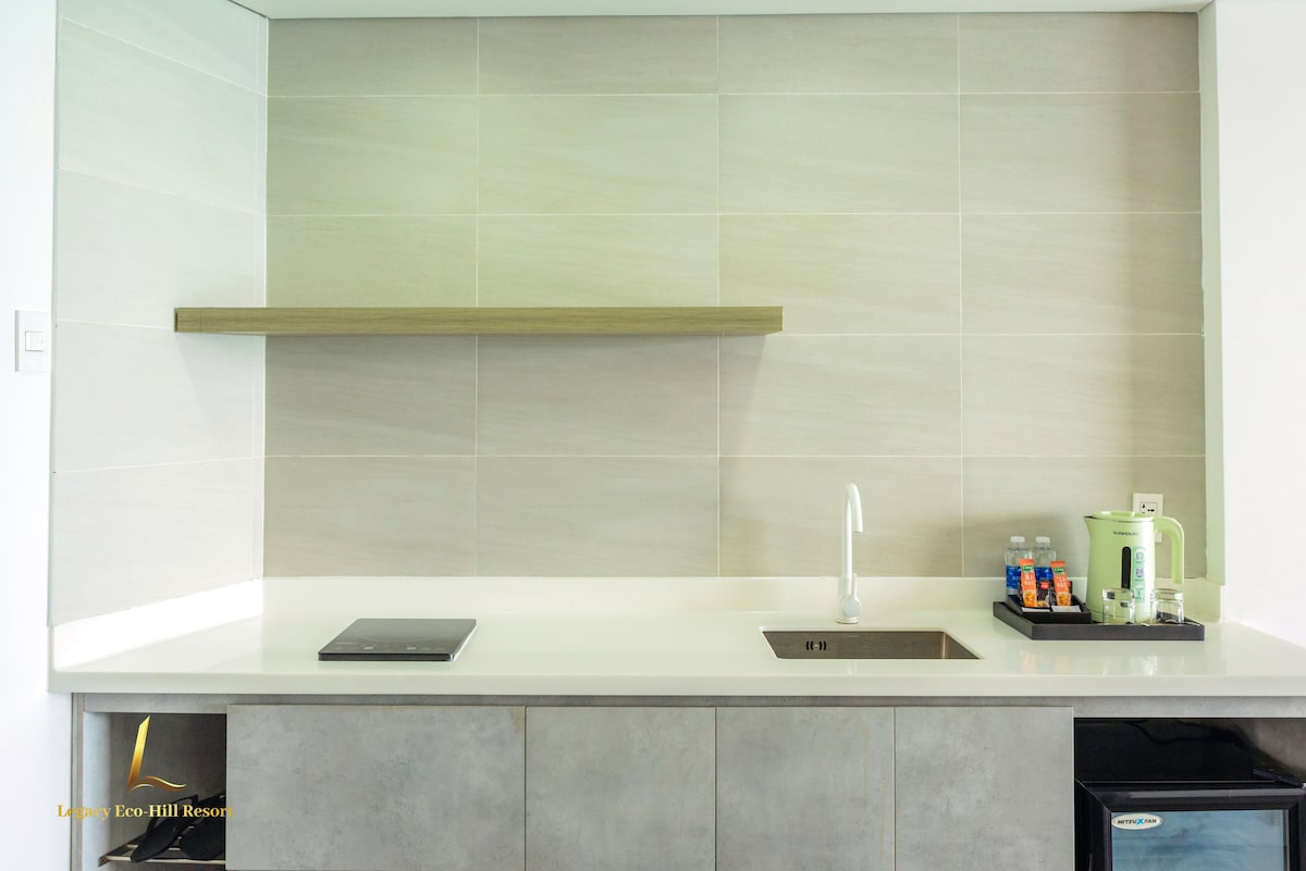 A modern kitchenette is depicted, featuring a sleek countertop with a small sink and an induction cooktop. Above, a wooden shelf provides space for storage. A compact refrigerator is positioned nearby, alongside a kettle and complimentary bottled water, enhancing the functional design.