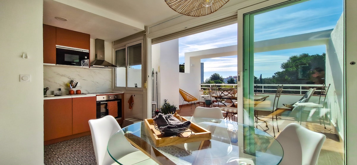 An open-plan dining area features a glass table accompanied by white chairs. A well-equipped kitchen is visible in the background, with modern cabinetry and appliances. Sliding glass doors lead to a sunlit terrace, showcasing a view of greenery and blue skies.