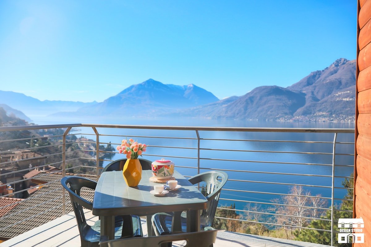 A balcony table set for four overlooks a stunning 180-degree view of Lake Como. A small vase with flowers rests on the table, while the scenic mountains provide a tranquil backdrop under clear blue skies.