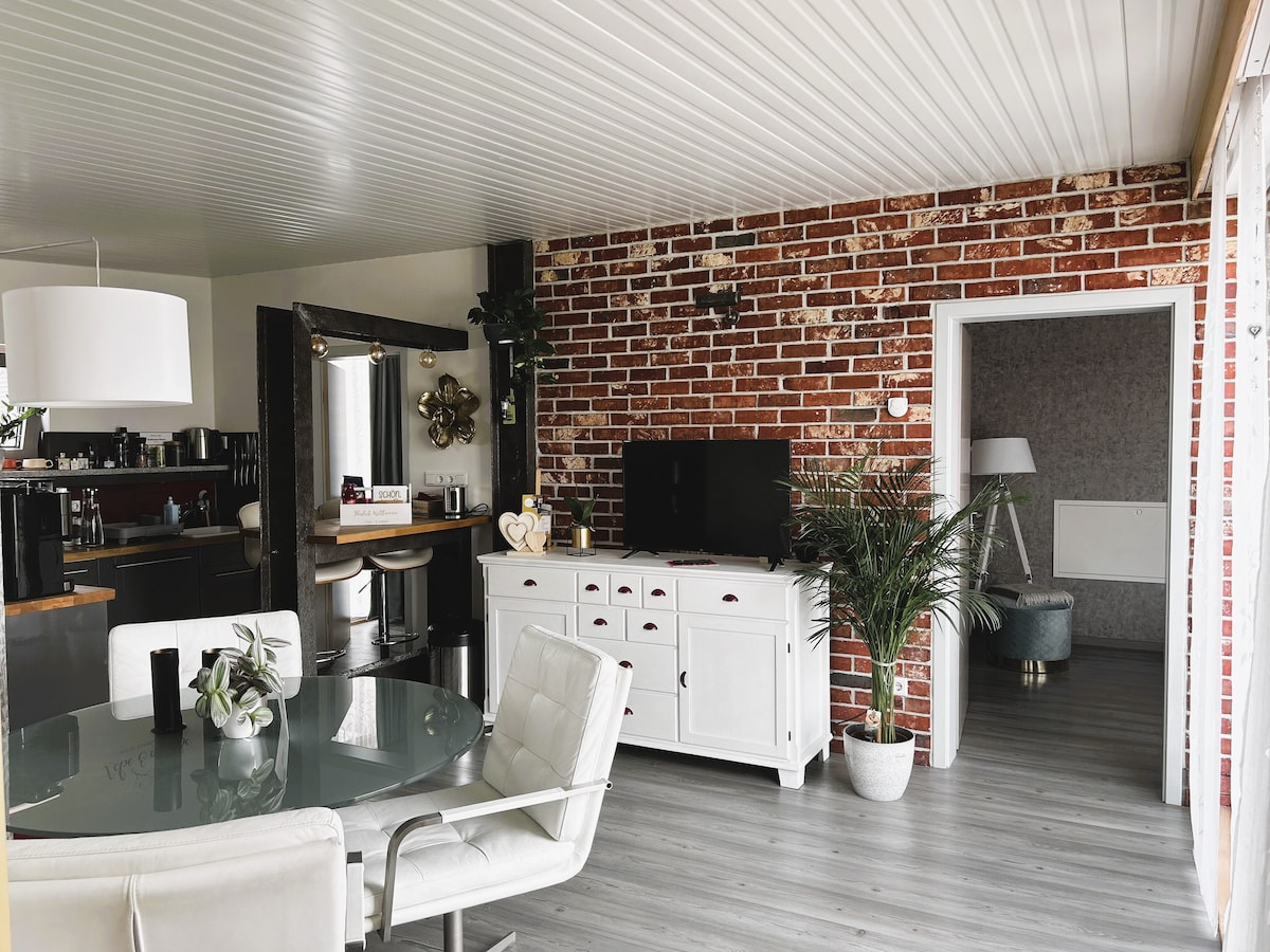 A bright living space features a circular glass dining table surrounded by white chairs. A white cabinet stands against a exposed brick wall, while a television is displayed on a modern stand. A doorway leads to another room, and greenery is present throughout the area.