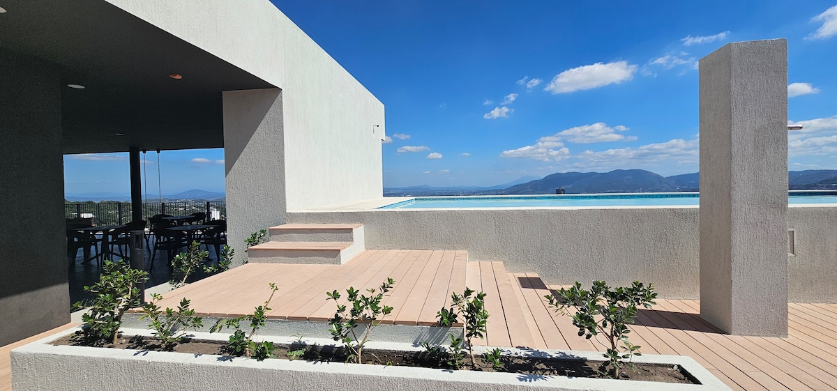 An outdoor terrace is presented with wooden decking and modern architecture. A rooftop pool can be seen in the background, framed by landscaped greenery. The blue sky with scattered clouds provides a bright backdrop, while seating areas are visible in the distance.