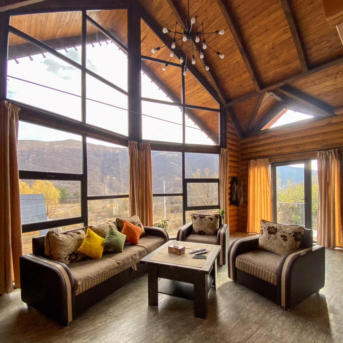 A spacious living area features high wooden ceilings and large windows that frame scenic views of the surrounding landscape. Comfortable seating is arranged around a central coffee table, complemented by colorful cushions. Natural light floods the space through the expansive glass panels.
