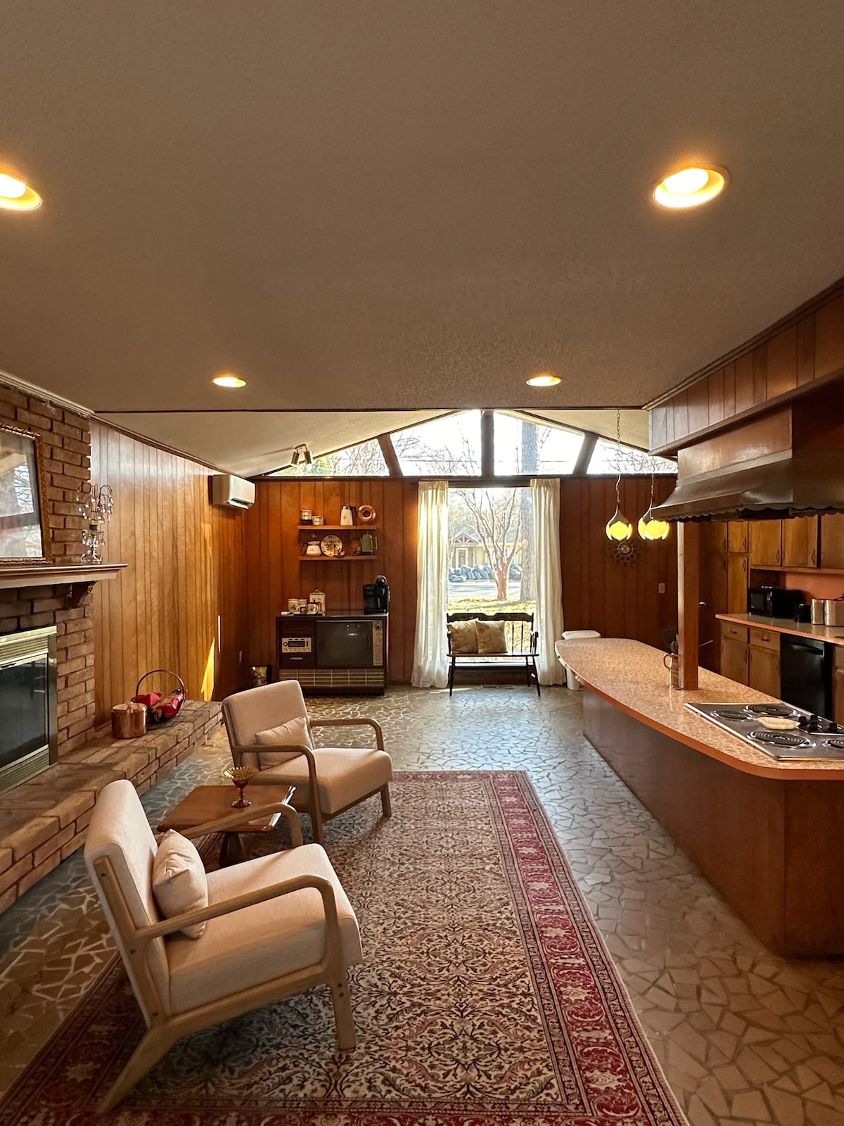 A spacious living area features a wooden accent wall and a stone fireplace. Natural light floods the room through large windows. Two light-colored chairs are placed on a patterned rug, with a kitchen area showcasing a long countertop and modern appliances in the background.