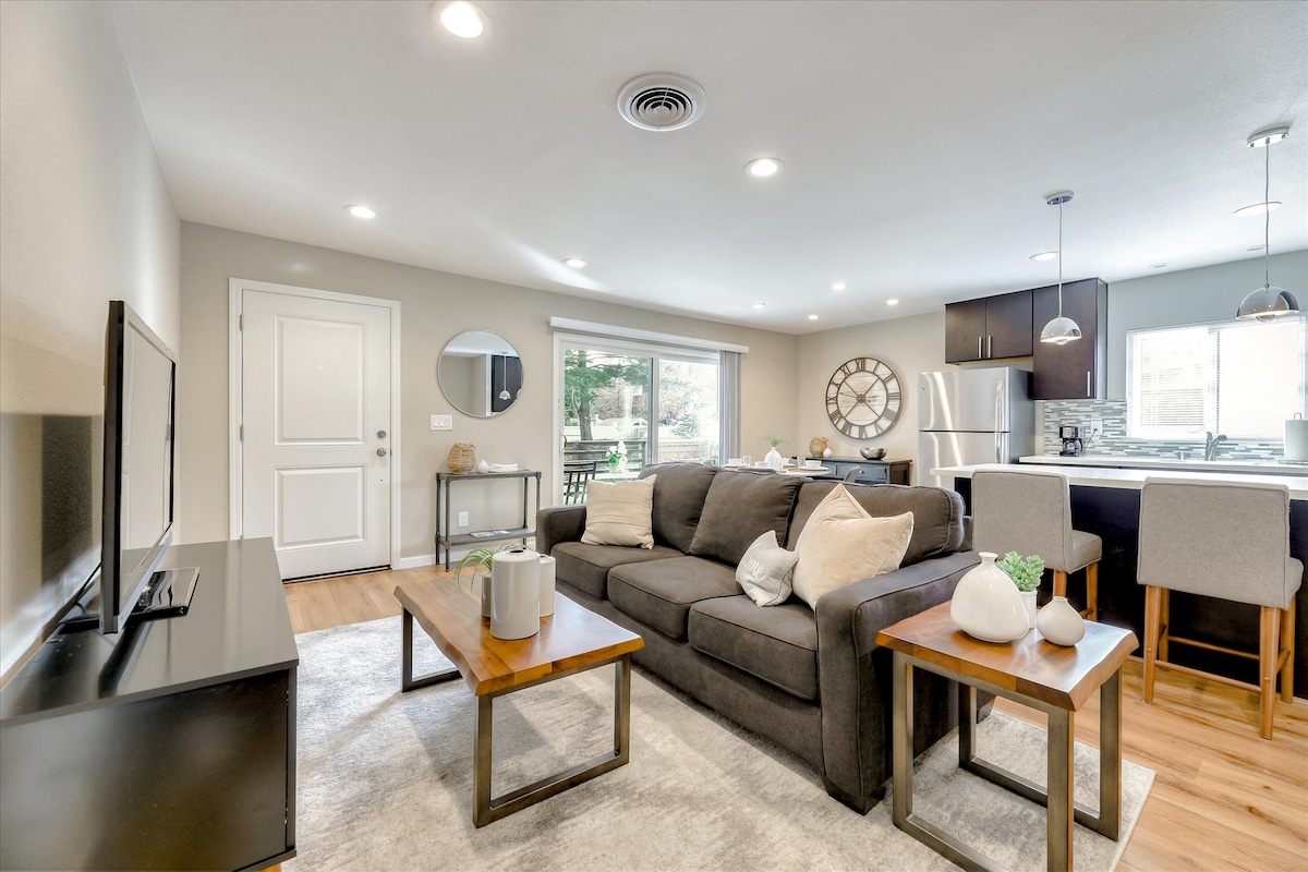 A modern living space features a comfortable sofa with wood accent tables. Natural light floods in through large windows, illuminating the neutral color palette. An open layout connects the living area with a well-equipped kitchen, highlighted by stylish cabinetry and contemporary lighting fixtures.