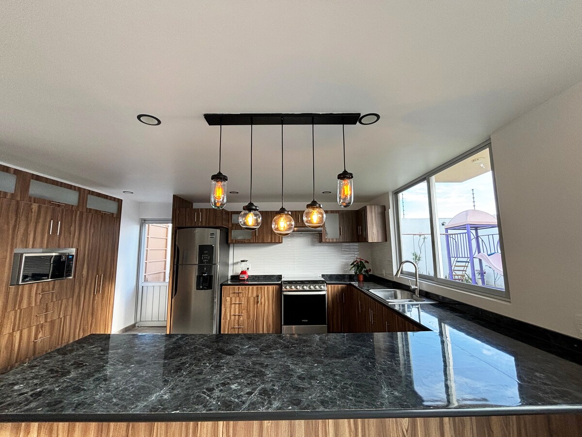 A well-lit kitchen features a large dark countertop with a modern stove and stainless steel refrigerator. Overhead, decorative pendant lights provide warm illumination. Large windows allow natural light to fill the space, and sleek wooden cabinetry complements the overall design.