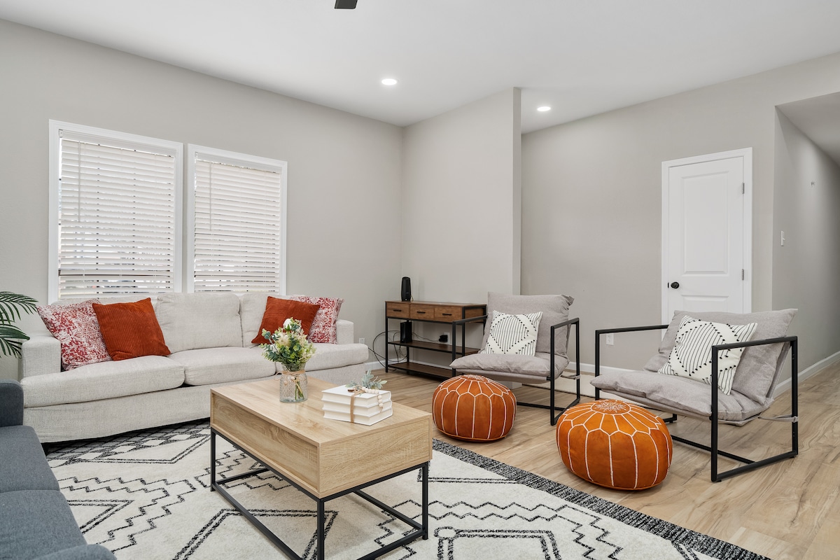 A spacious living area features a light-colored sofa adorned with orange and patterned cushions. Two inviting armchairs with geometric designs are positioned around a wooden coffee table. A patterned area rug complements the room's neutral walls, while natural light streams through the window coverings.