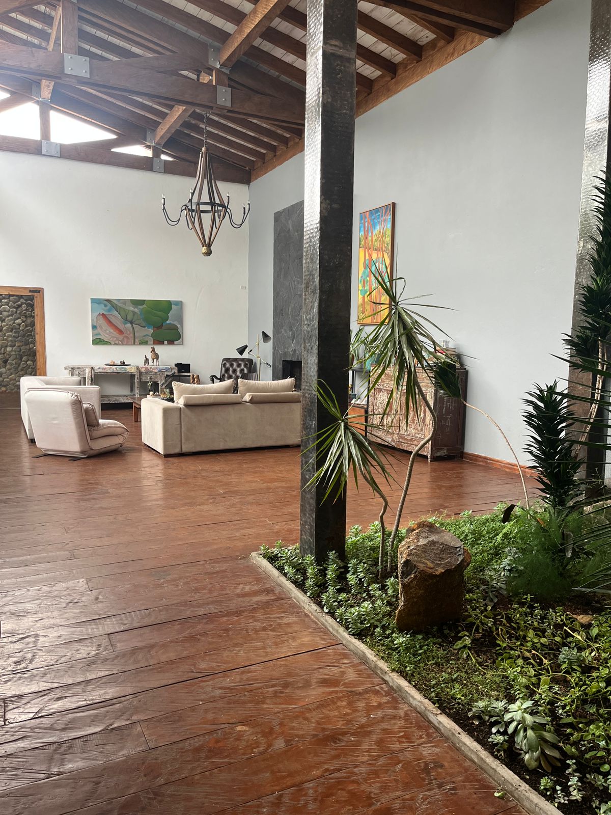 An open living area features a high ceiling with wooden beams. Natural light filters through large windows, illuminating the spacious seating arrangement and a modern chandelier. A vertical garden with plants and a small rock adds a touch of greenery to the space.