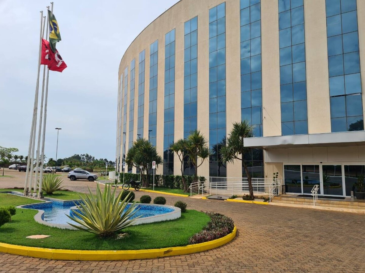 The entrance of the building showcases large glass windows and a curved façade. A small fountain is situated in a landscaped area, featuring green plants and decorative features. Flags are displayed on poles, and the surrounding parking area is visible in the background.