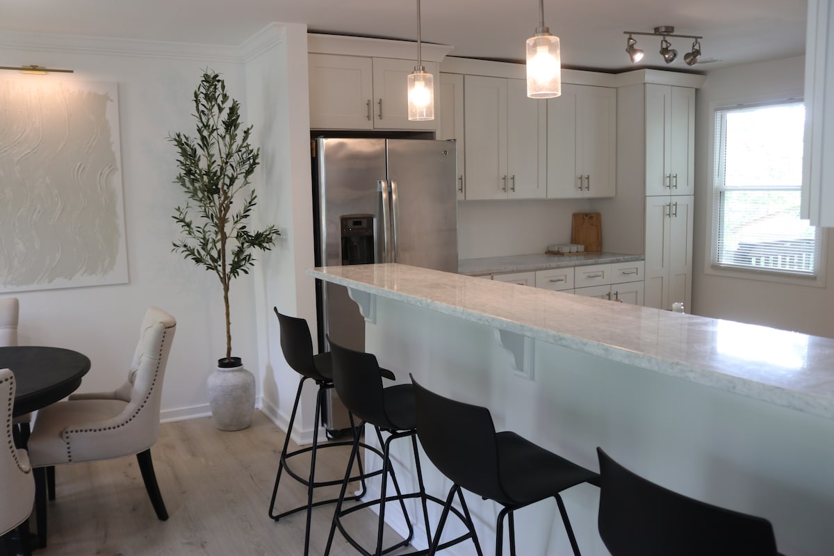 A modern kitchen features sleek white cabinetry and stainless steel appliances. A long countertop with four black bar stools offers seating. Natural light fills the space through a window, while a potted plant provides a touch of greenery.