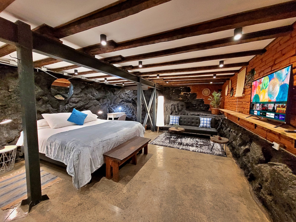 An inviting room features a spacious bed adorned with a light blanket and an accent pillow. A sleek black couch complements the rustic stone wall, while a television is mounted above a wooden shelf. Modern lighting fixtures illuminate the room, highlighting its unique character.