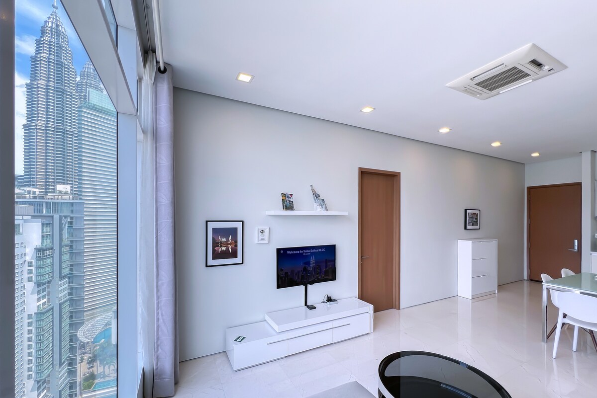 A modern living room features a wall-mounted TV under a sleek white cabinet. Large windows provide a view of the nearby Petronas Towers, letting in natural light. Light-colored tile flooring complements the minimalist decor, while a door leads to additional spaces.