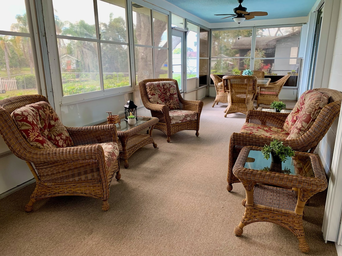 A spacious covered patio is furnished with comfortable wicker seating featuring floral cushions. Two small tables are placed between the chairs, and potted plants add touches of greenery. Large windows allow natural light to illuminate the space while offering views of the surrounding landscape.