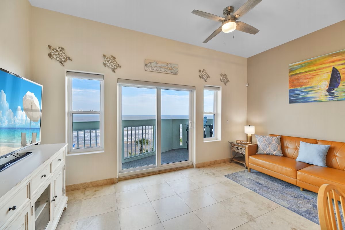 A bright living area features a large window with ocean views and a sliding glass door leading to the balcony. A comfortable orange sofa and a light wooden side table are positioned nearby. A ceiling fan and a flat-screen television add to the inviting ambiance.
