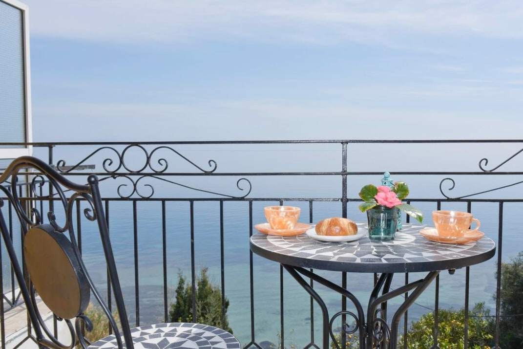 A balcony table set with two decorative cups and a croissant rests against a backdrop of the Ionian Sea. A small vase with fresh flowers adds a touch of color. The railing offers an unobstructed view of the tranquil waters and sky.