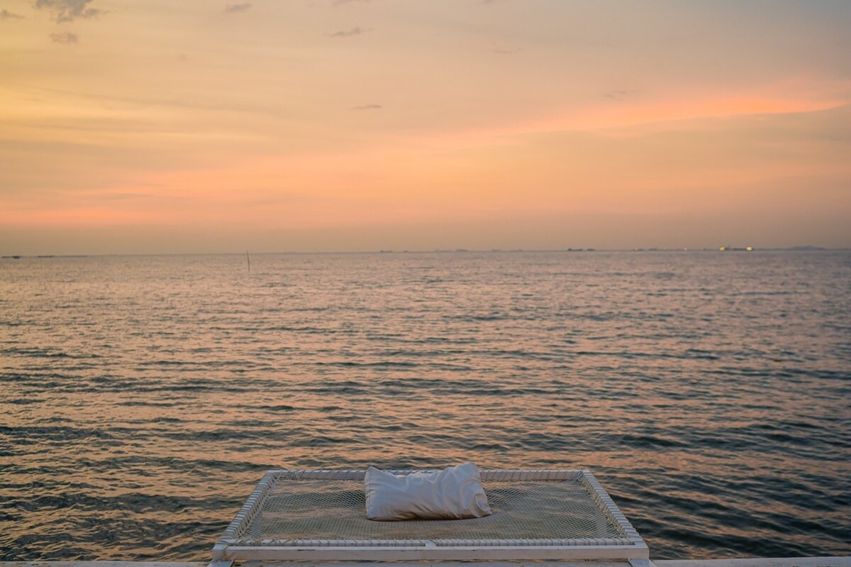 A serene view of the ocean stretches to the horizon, reflecting soft pastel hues of sunset. A white cushion is placed centered on a metal lounge frame, offering a comfortable spot to enjoy the tranquil waters and sky.