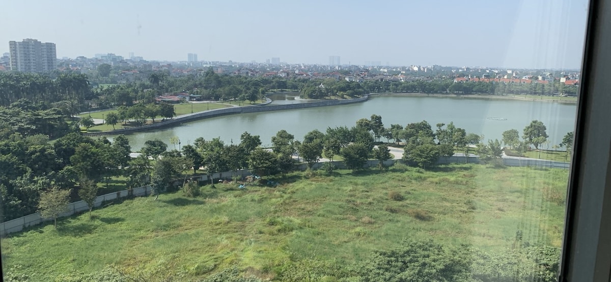 A panoramic view of a serene lakeside landscape is presented, featuring lush greenery and a calm body of water. The surrounding area includes park pathways and distant city buildings, providing a sense of tranquility amidst urban life.
