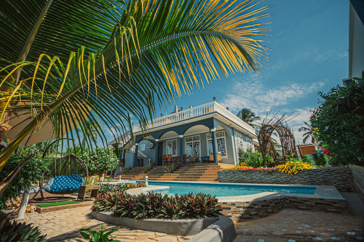 A vibrant garden surrounds a tranquil pool area, highlighted by lush tropical plants and colorful flowers. The villa, featuring a spacious porch with seating, offers a welcoming ambiance against a clear blue sky. A shaded swing adds to the serene outdoor space.