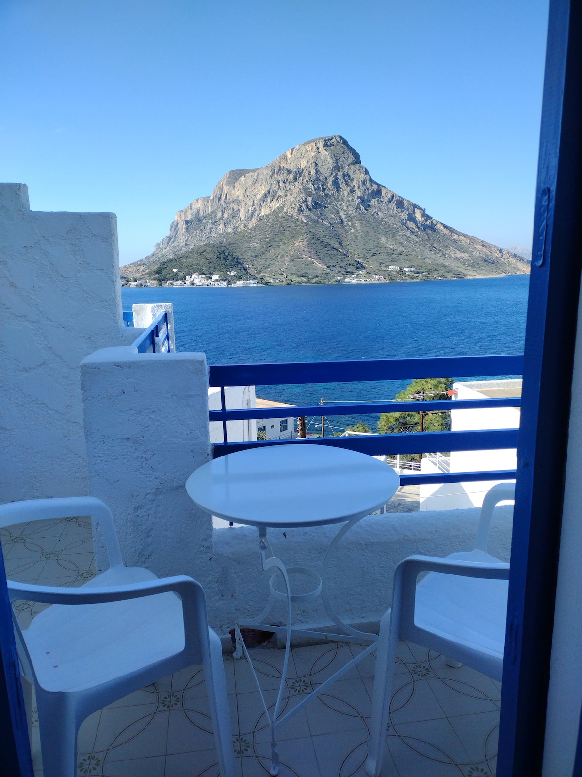 A balcony is shown with a round table and two white chairs. A stunning view of a mountainous landscape is visible in the background, overlooking the blue waters of the sea under a clear sky.