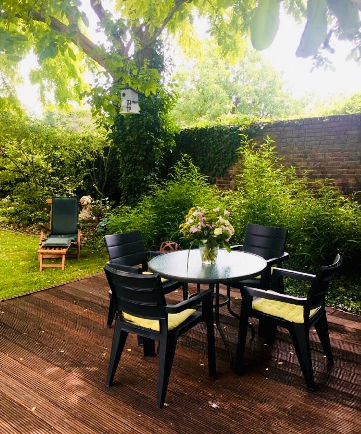 A wooden terrace features a round glass table surrounded by five chairs, each with yellow seat cushions. Lush greenery surrounds the area, including a tall tree providing shade. A bird feeder is visible hanging from a branch, enhancing the garden's natural ambiance.