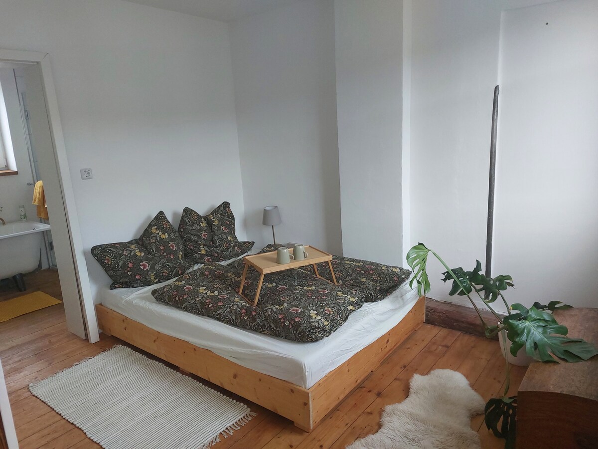 A cozy sleeping area features a spacious wooden bed adorned with a patterned duvet and multiple pillows. A small bedside table holds two cups, and a lamp provides soft illumination. A plant and a light-colored area rug enhance the tranquil environment.