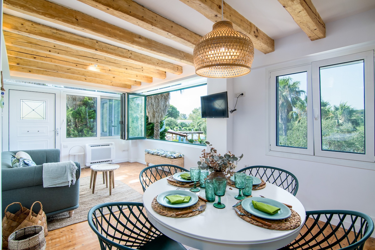 An open-plan living space features a circular dining table set with green dishware and transparent glasses. A comfortable gray sofa is positioned nearby, complemented by wooden beams and large windows offering scenic views of the surrounding greenery.