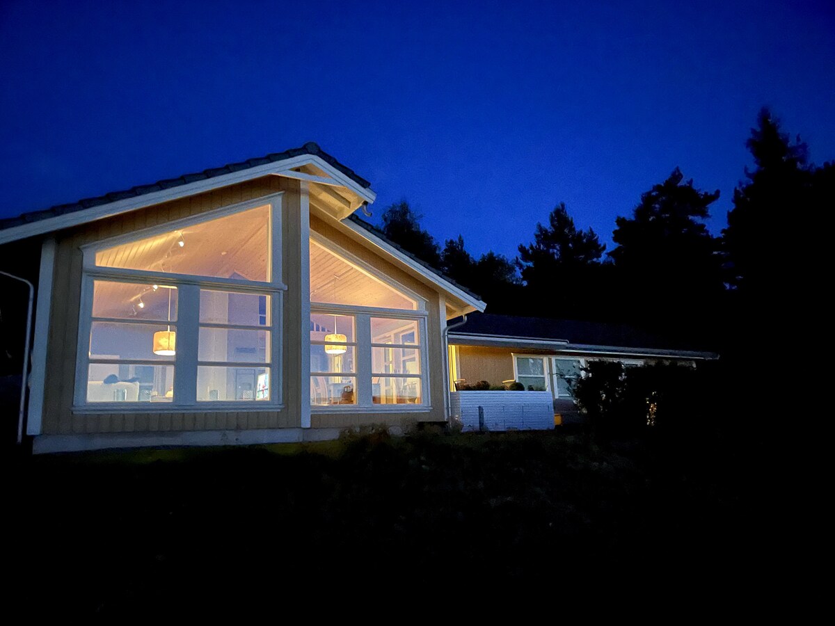 The exterior of the cabin is shown against a night sky, with warm light emanating from large windows. The structure's modern design is highlighted by a sloped roof and natural surroundings, creating a welcoming atmosphere.