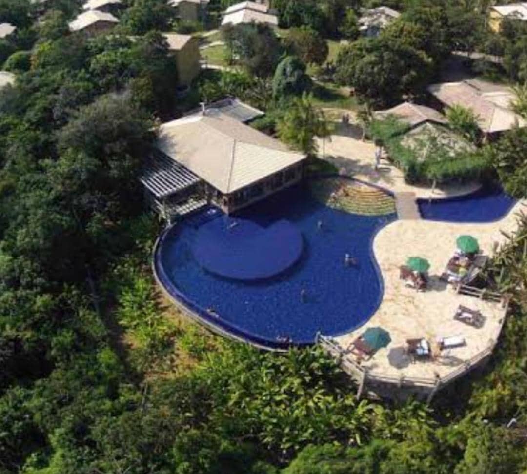 An aerial view captures a large, inviting pool surrounded by lush greenery. The pool features a unique shape with a central circular area. Comfortable lounge chairs are placed around the pool area, and shaded umbrellas provide spots for relaxation.